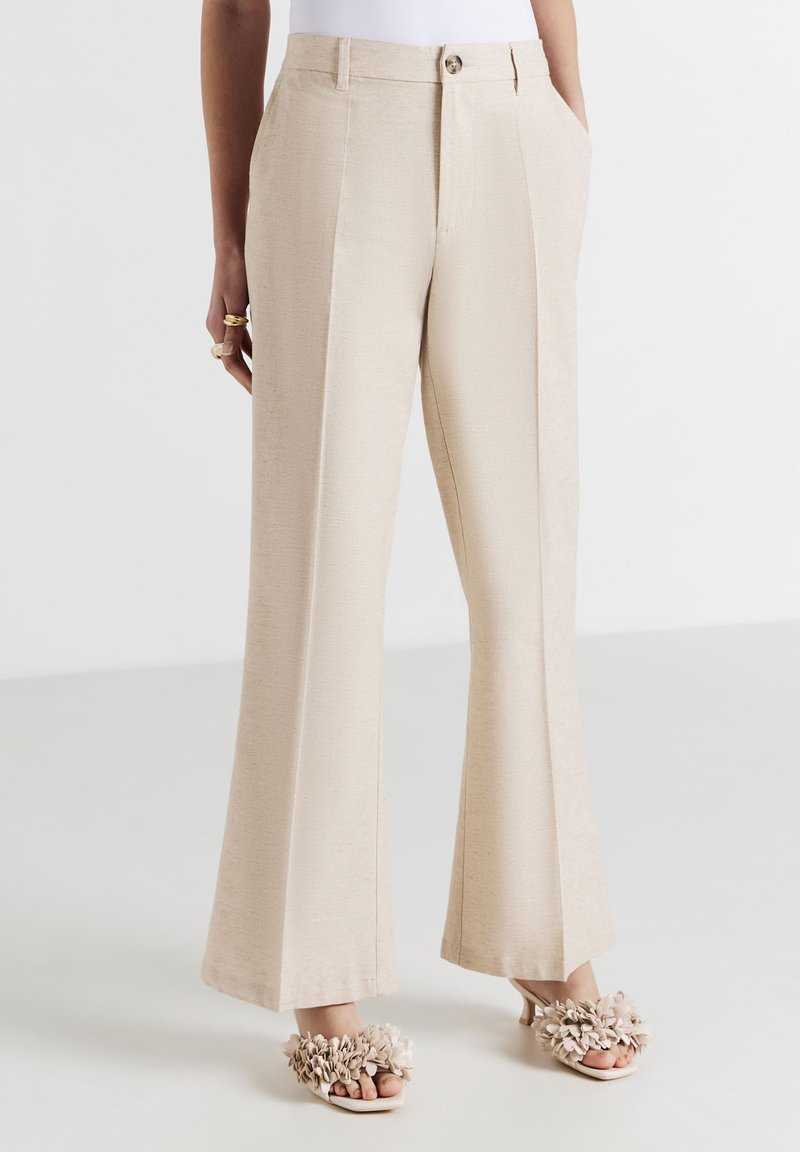 Beige hochgeschnittene weit geschnittene Hosen mit vorderen Falten, seitlichen Taschen, Knopf- und Reißverschluss, getragen mit beige blumengeschmückten offenen High Heels.