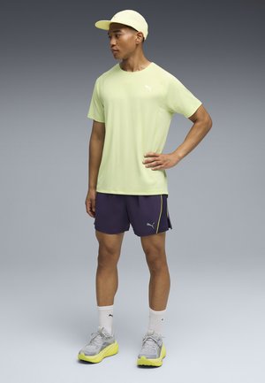 Camiseta deportiva de color amarillo claro, pantalones cortos morados con detalles en amarillo, calcetines blancos, zapatillas de deporte grises con suelas amarillas y una gorra a juego.