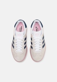 Zapatillas Adidas con un upper de gamuza color beige, rayas azul marino, cordones blancos y una plantilla de color rosa suave. Punta redonda y suela de goma.