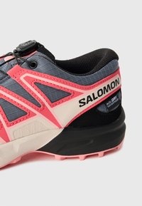 Trail running shoe featuring a gray textile upper with pink accents, a black heel, and a textured rubber sole. Includes adjustable lacing system.