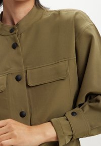 Vista ravvicinata di una persona che indossa una giacca verde oliva con bottoni, dotata di tasche a pattina e polsini con bottoni.