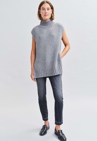 Maglione senza maniche grigio a maglia con collo alto, caratterizzato da un orlo a coste e tasche laterali, abbinato a jeans slim scuri e mocassini neri.