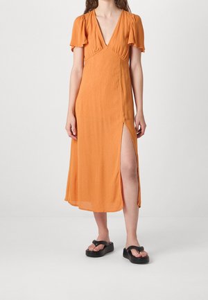 Mujer con un vestido midi naranja con mangas acampanadas y abertura alta en el muslo, combinado con sandalias negras de plataforma tipo chanclas.