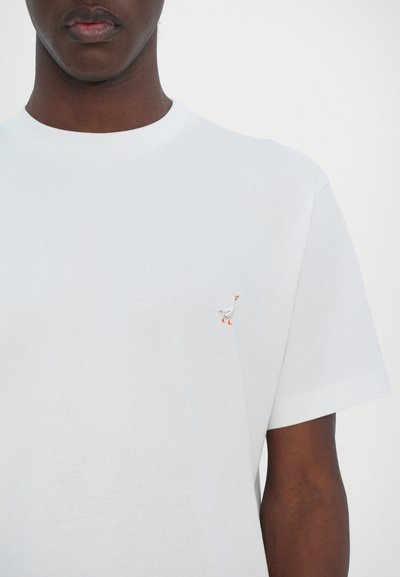 T-shirt en coton blanc avec un col rond et des manches courtes, ornée d'un petit motif de canard brodé en gris et orange sur la poitrine.