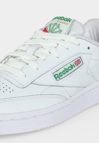 Biele tenisky Reebok Classic s bielymi šnúrkami, zeleno-červeným logom a textúrovanou gumovou podrážkou viditeľnou na svetlom pozadí.