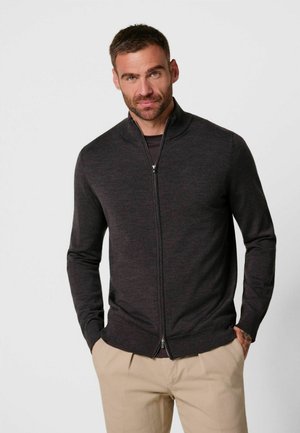 Uomo con capelli corti e barba che indossa un maglione grigio scuro con zip e pantaloni beige, in piedi con le mani nelle tasche su uno sfondo semplice.