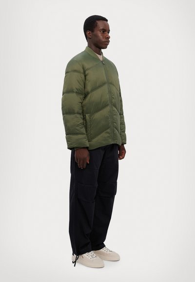 Veste matelassée verte avec un design en chevrons, fermeture éclair à l'avant et col côtelé. Associée à un pantalon cargo bleu marine et des baskets blanches.