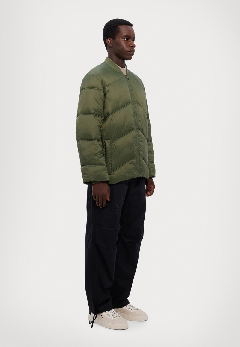 Veste matelassée verte avec un design en chevrons, fermeture éclair à l'avant et col côtelé. Associée à un pantalon cargo bleu marine et des baskets blanches.