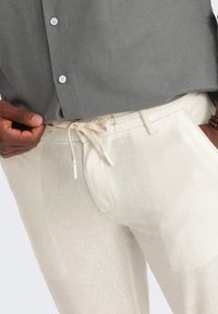 Pantalon en lin beige clair avec une taille élastique à cordon et des poches avant, associé à une chemise grise à boutons.