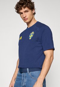 Κοντομάνικο μπλε σκούρο μπλουζάκι με το έμβλημα της Σουηδίας και κίτρινες λεπτομέρειες. Διαθέτει στρογγυλή λαιμόκοψη και υφασμάτινες επενδύσεις στους ώμους. Φοριέται με μπλε τζιν παντελόνι.