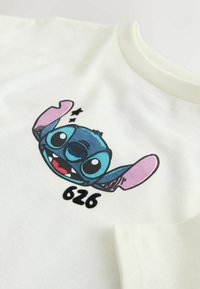 T-shirt en coton blanc avec un imprimé coloré de personnage de dessin animé représentant un extraterrestre bleu aux oreilles roses et contour noir, étiqueté "626" en dessous.