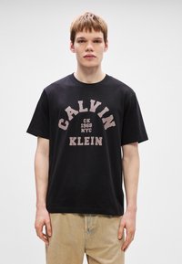 Črna bombažna majica z osrednjo grafiko "CALVIN KLEIN CK 1968 NYC" v svetlo roza barvi. Kratki rokavi in okrogla ovratnika.