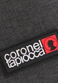 Fondo de tela negra con un parche rectangular de logo. El parche presenta "coronel tapiocca" en texto blanco y un diseño de silueta roja.