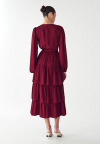 Burgundowa sukienka tiered z długimi rękawami, ze zgromadzonym w pasie materiałem, zwiewnym krojem i miękką fakturą, w zestawieniu z jasnymi sandałami na obcasie.