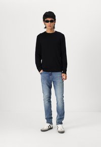 Camisa negra de manga larga, jeans azules claros y zapatillas blancas con acentos grises. Modelo usando gafas de sol negras frente a un fondo liso.