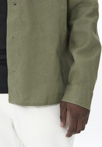 Camisa verde oliva de botonadura con una tela texturizada, que presenta un corte relajado. Los puños están doblados y se puede ver una capa inferior negra con relieve.