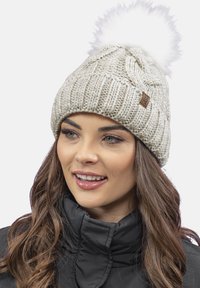 Beanie lavorato a maglia di colore beige con un pompon in pelliccia sintetica bianca sulla cima e un motivo testurizzato. Presenta un piccolo etichetta in pelle marrone sul lato.