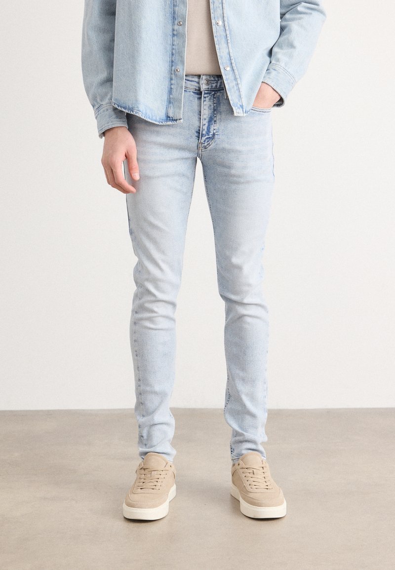 Jeans de mezclilla azul claro con corte slim, con desvanecimiento mínimo y detalles de costura. Combinados con zapatillas beige sobre un fondo neutro.