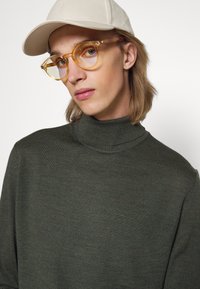 Kaki turtleneck genser med fin tekstur, kombinert med transparente gule katteøye-briller og en beige baseballcap.