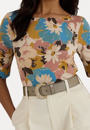Vrouw draagt een meerkleurige bloemenblouse met blauw, mosterd en roze tinten, ingestopt in een hooggetailleerde crèmekleurige broek met een metalen riem.