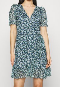 Květinové šaty s hlubokým výstřihem do V, krátkými nabíranými rukávy a rozšířeným spodním lemováním. Vyrobené z lehkého materiálu s modrými a zelenými květinovými vzory.