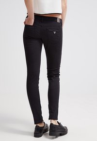 Jeans ajustados negros con textura suave, bolsillos traseros y un pequeño emblema en la cintura. Combinados con zapatos negros con suelas texturizadas.