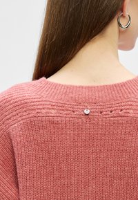 Pletena majica v mehki roza barvi z rebrastim ovratnikom, ki ima dekorativne svetle luknje in majhno kovinsko gumbico na hrbtni strani.