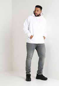 Fehér kapucnis pulóver, kenguruzsebbel, szürke slim fit farmernadrággal és fekete sportcipővel párosítva, világos háttér előtt.