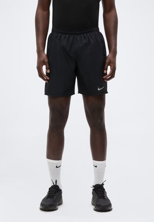 Mand iført sorte atletikshorts med Nike-logo, sorte sneakers og hvide Nike-strømper står mod en ensfarvet baggrund.