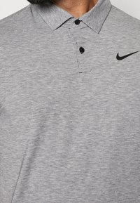 Graues Poloshirt aus strukturiertem Stoff, mit flachem Kragen, zwei Knöpfen und einem schwarzen Nike-Logo auf der linken Brust.