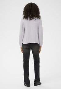 Lys lavendelbluse med lange pufærmer, løs pasform og tekstureret stof, parret med mørke jeans og sorte sko. Bagview.