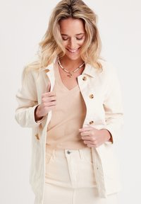 Veste légère beige à boutons avec une coupe décontractée, comportant deux poches avant et des boutons marron. Portée sur un haut léger beige.