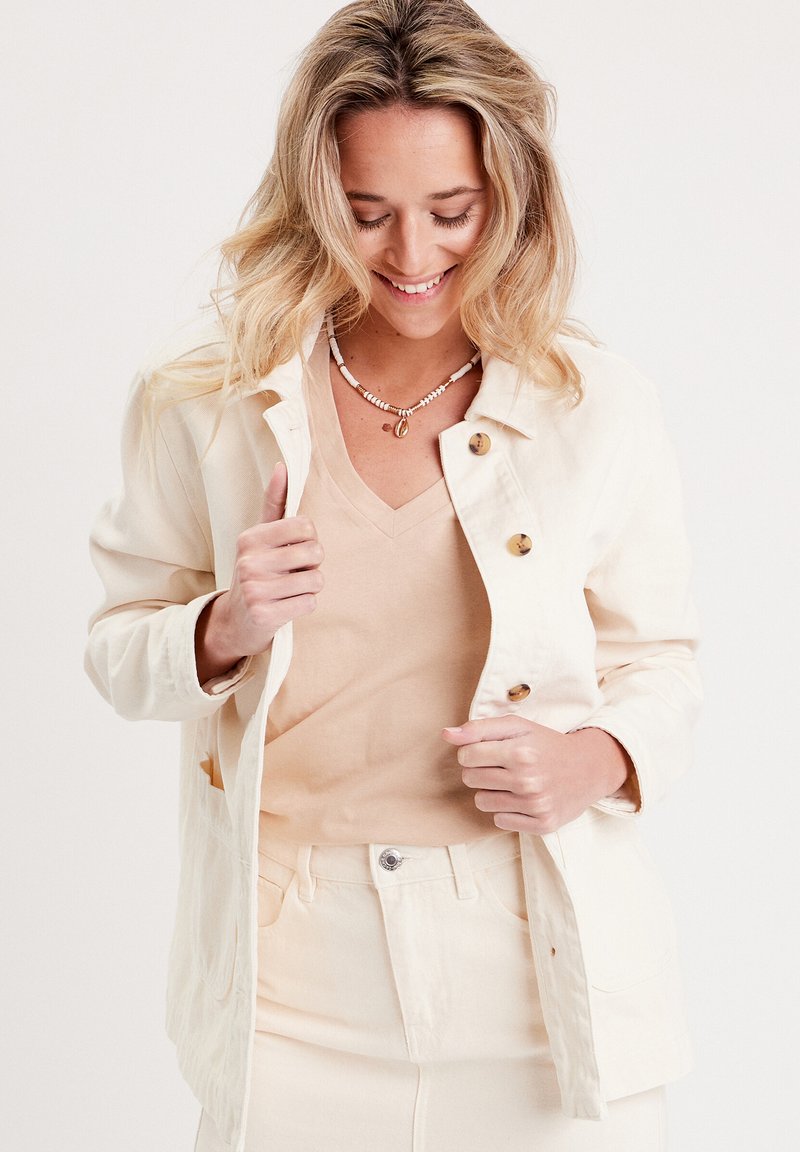 Veste légère beige à boutons avec une coupe décontractée, comportant deux poches avant et des boutons marron. Portée sur un haut léger beige.