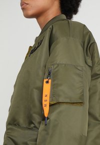 Veste bomber vert olive avec une poche zippée et une étiquette orange sur la manche. Tissu lisse et brillant avec des broderies sur le devant.