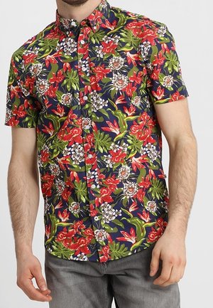 Homme portant une chemise à manches courtes à boutons avec un motif floral coloré rouge, vert et blanc sur un fond bleu foncé.