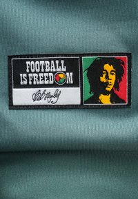 Lapp på blått stoff med teksten "FOOTBALL IS FREEDOM", farget fotballlogo, signatur og stilisert portrett på rød, gul, grønn bakgrunn.