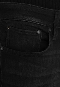 Mustat farkut, joissa on etutasku metallisten niittien kanssa ja hienovarainen rakenne. Vyötäröllä on vaakasuora kudottu kuvio.