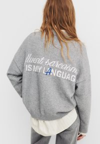 Maglione grigio lavorato a maglia con vestibilità ampia, con la scritta "sarcasm IS MY LANGUAGE" in bianco e un accento blu sulla lettera A. Bordo e polsini a coste.