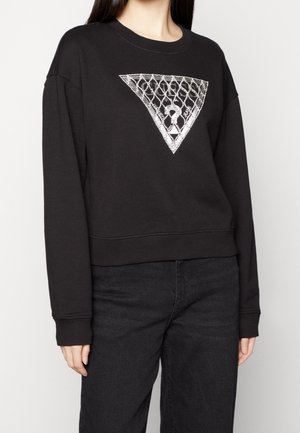Femme portant un sweat-shirt noir avec un motif triangulaire en maillons de chaîne argentés et un point d'interrogation au centre, associé à un pantalon noir.