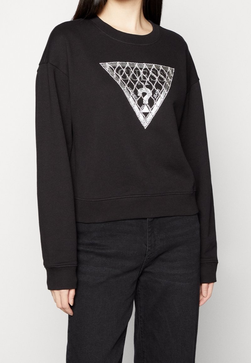 Femme portant un sweat-shirt noir avec un motif triangulaire en maillons de chaîne argentés et un point d'interrogation au centre, associé à un pantalon noir.