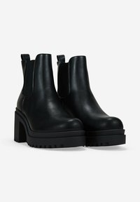 Botas de cuero negro hasta el tobillo con un tacón grueso y una suela de goma con estrías. Incluyen paneles laterales elásticos y lengüetas para facilitar el calzado.
