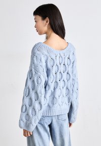 Gaiši zils adīts džemperis ar brīvu piegriezumu, ar lieliem ovāliem izgriezumiem un ribinātām aprocēm. Aizmugurē atvērts kakla dizains, apvienots ar gaišiem džinsa auduma izstrādājumiem.