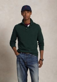 Žalias ilgomis rankovėmis polo marškinėliai su apykakle, užsidėti virš bordo spalvos marškinėlių, derinami su nuplyšusiais mėlynais džinsais ir jūrų spalvos beisbolo kepure.