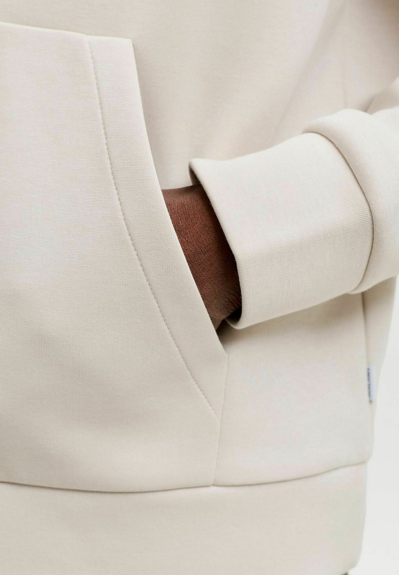 Main à la peau foncée partiellement dans la poche avant d'un sweat-shirt couleur crème avec une côte visible au niveau du poignet.