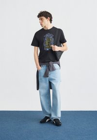 Νεαρός άντρας με μαύρο γραφικό μπλουζάκι, ανοιχτό μπλε τζιν και μαύρα αθλητικά παπούτσια, κρατώντας μια μαύρη τσάντα ώμου, στέκεται σε μπλε χαλί.