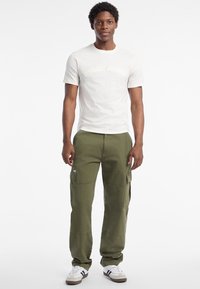 Helle Cremefarbene Herren-T-Shirt mit geprägt Logo, kombiniert mit olivgrünen Cargo-Hosen mit seitlichen Taschen und umgeschlagenen Säumen.