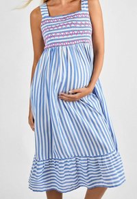 Mujer embarazada vistiendo un vestido sin mangas a rayas azul y blanco con bordados en zigzag rosa en el pecho, sosteniendo su barriga.