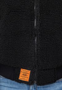 Giacca in pile nera con tessuto testurizzato e morbido, chiusura con zip e etichetta arancione nell'orlo. Presenta polsini e vita a costine.