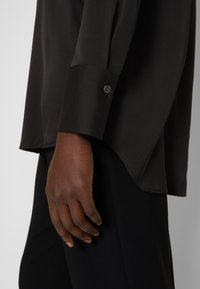 Main à la peau foncée reposant sur le côté portant une chemise noire à manches longues avec des poignets boutonnés et un pantalon noir sur fond gris.