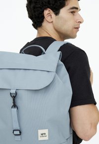 Ljusblå ryggsäck med lockstängning, med en framficka, justerbara bälten och en etikett på den nedre framsidan. Tillverkad av texturerad tyg.